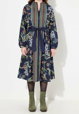 Femme portant une robe midi bleu marine à motif floral avec des panneaux verticaux, des manches longues, un nœud à la taille, des collants noirs et des bottines vert olive.