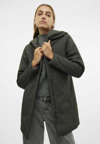Chaqueta impermeable de color verde oliva, con capucha, cremallera frontal y botones a presión, llevada sobre un suéter de punto gris y jeans holgados.