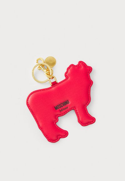 Breloc din piele roșie în formă de vacă, cu accesorii din metal aurit și logo-ul embosat "Moschino Milano". Textură netedă și margini definite.