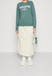 Felpa verde con scritta "HOLLISTER California", gonna midi color crema con tasche cargo, sneakers bianche chunky e borsa riflettente.