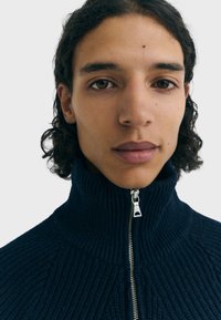 Marineblauwe sweater met ritssluiting, geribbelde textuur, hoge kraag en zilveren ritstrekker. De stof lijkt dik en warm.