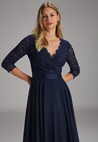 Laivansinis mekko, jossa on pitsinen bodysuit, jossa on paljetteja, aaltoileva kaula-aukko ja kolmannes hihasta. Liikkuva hame yksinkertaisesta kankaasta.