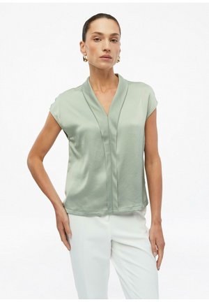 Femme aux cheveux lissés en arrière portant un chemisier vert clair à manches courtes et un pantalon blanc, posant avec une main sur la hanche sur fond blanc.