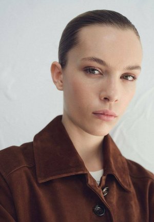 Braune Wildlederjacke mit großem Kragen und Knopfverschluss. Glatte Textur mit entspanntem Sitz, getragen über einem weißen Oberteil vor hellem Hintergrund.