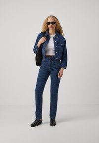 Sada džínové bundy a kalhot v tmavě modré barvě s geometrickým vzorem. Bílé crop top a černé doplňky dotvářejí celý outfit. Černé vysoké podpatky.