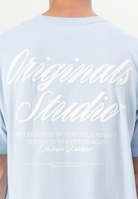 Svijetloplava majica s kratkim rukavima, s bijelim natpisom na leđima. Natpis uključuje "Originals Studio" i detalje dizajna u različitim fontovima.