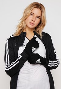 Zwarte sporthandschoenen met wit Adidas-logo; zacht, passend materiaal; gedragen met een zwarte jas met drie witte strepen.