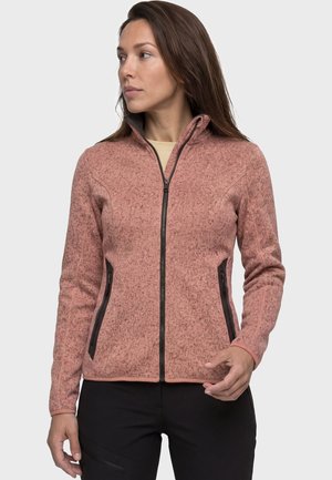 Frau trägt eine pinkfarbene Fleecejacke mit Reißverschluss und schwarzen Reißverschlusstaschen, steht mit leicht erhobener linker Hand und schaut nach links.