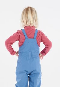 Blå vattentäta overaller med justerbara axelremmar, bärs över en röd fleece-tröja. Slät yta, elastiskt midjeband, logotyp på baksidan.