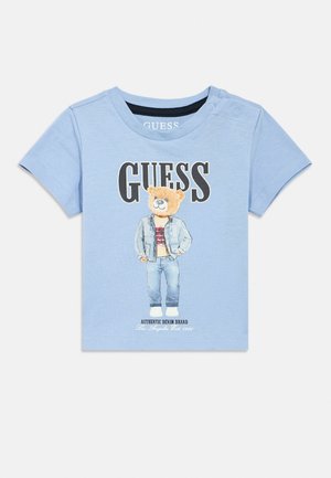 Lyseblå t-shirt til småbørn med en bjørn iført denimtøj og ordet "GUESS" i store bogstaver ovenover.