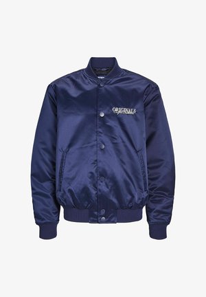 Tengerészkék selyem bomber dzseki fekete hoodiéval, világoskék, buggyos farmernadrággal és fehér sportcipővel párosítva. Logó részletek jellemzik.