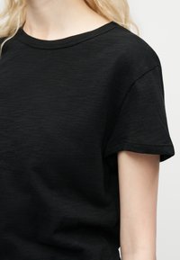 Personne blonde portant un t-shirt noir uni à manches courtes avec un col rond sur un fond clair.