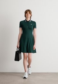 Zielona sukienka polo z krótkimi rękawami, kołnierzykiem i białymi paskami. Stylizowana z czarną torebką i białymi trampkami. Gładka tekstura.