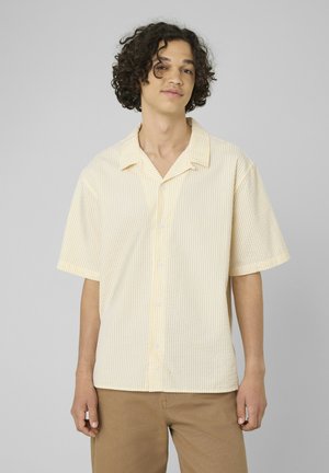 Jeune homme aux cheveux bouclés portant une chemise à manches courtes rayée jaune clair à boutons et un pantalon marron sur un fond uni.