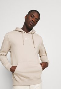 Beige kapuce ar pelēko džemperi ar kabatu un auklu. Iespējo nelielu logotipu uz krūtīm un - kabatu uz rokas. Gluda tekstūra.