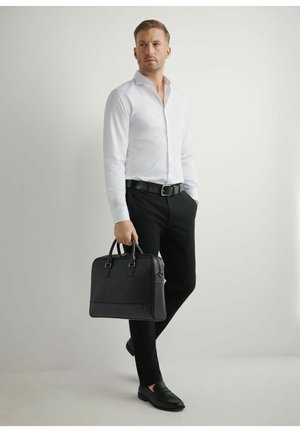 Mężczyzna w białej koszuli i czarnych spodniach, trzymający czarną teczkę, stojący na jednolitym jasnym tle.