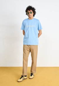 Light blue t-shirt with “carhartt.” text in beige, paired with tan pants and yellow/black shoes. Model wears green sunglasses.