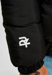 Manșonul unei jachete negre căptușite prezintă un logo alb. Materialul este neted, cu manșete elasticizate, evidențiind un design voluminos.