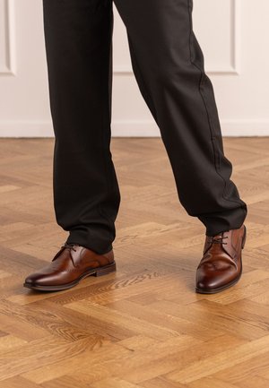 Brown leather dress shoes with brogue detailing worn with black trousers on light wooden parquet floor.