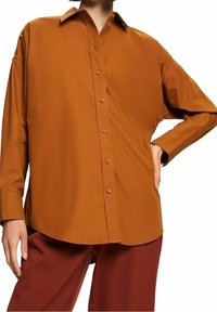 Camisa de botón de manga larga color óxido con cuello, de corte relajado y textura suave. Presenta botones en la parte delantera y un dobladillo curvado.