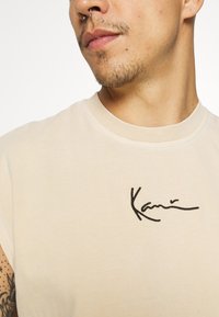 Beige bomull t-skjorte med korte ermer, med svart brodert signaturdetalj på brystet og rund halslinning.