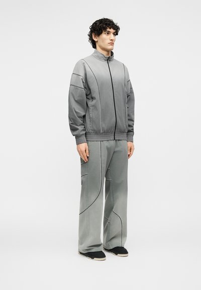 Νεαρός άνδρας όρθιος, φοράει ένα ταιριαστό ανοιχτό γκρι σακάκι και παντελόνι με φαρδιά πόδια, με μαύρες λεπτομέρειες περιγράμματος και μαύρα παπούτσια.