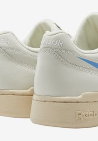 Sneakers bianchi in pelle con un design a spunta blu chiaro, una suola spessa color crema e il marchio "Reebok" in rilievo sul tallone.