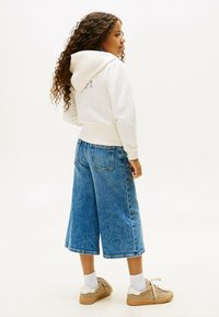 Biała bluza z kapturem i sznurkami, niebieskie dżinsowe culottes oraz beżowe sneakersy. Kręcone włosy i swobodna postura, ukazujące szczegóły i tekstury odzieży.