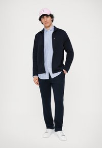 Casaco de fleece azul-marinho sobre uma camisa de botão às riscas azuis e brancas, combinado com calças escuras e ténis brancos. Acessorizado com uma capa rosa-claro.