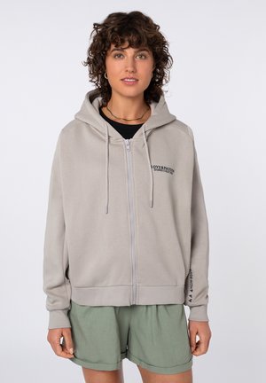 Junge Person mit kurzen lockigen Haaren, trägt einen hellgrauen Kapuzenpullover mit Reißverschluss und dem Schriftzug „LOVE & PASSION“ sowie grüne Shorts vor weißem Hintergrund.