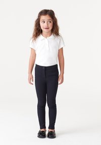 Polo shirt bianco a maniche corte, pantaloni aderenti blu navy con tasche frontali e scarpe nere con dettagli a fiocco. Tessuti semplici e lisci.