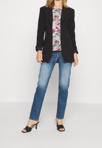 Schwarzer maßgeschneiderter Blazer über einem floral gemusterten Oberteil, kombiniert mit hellblauen Jeans und schwarzen Sandalen mit offenem Zeh. Glatte Textur und strukturierter Schnitt.