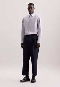 Camicia grigia chiara con bottoni e motivo sottile, abbinata a pantaloni a vita alta blu scuro. Scarpe eleganti nere completano il look.