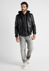 Schwarze Lederbomberjacke mit abnehmbarer Kapuze, kombiniert mit einem dunkelblauen Hemd, grauen Jeans und weißen Sneakern.