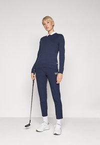 Tengerészkék V-nyakú pulóver, hosszú ujjú, tengerészkék szűkített nadrággal párosítva. A modell golf ütőt tart, fehér sportcipőt és zoknit visel.