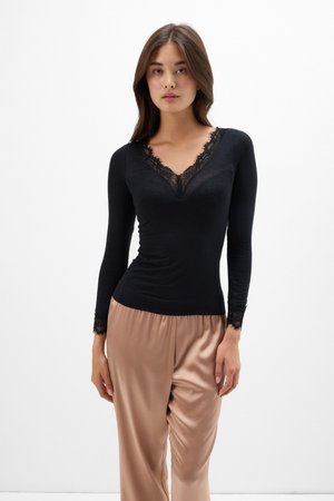 Mujer con top negro de manga larga con encaje en el escote y los puños, combinado con pantalones de satén beige sueltos, de pie frente a un fondo blanco.