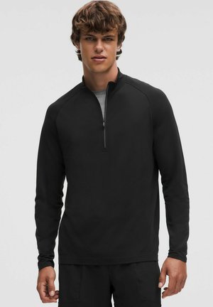 Zwarte lange mouwen zip-up top met een gestructureerde afwerking, gekenmerkt door een hoge kraag en raglanmouwen, gelaagd over een lichtgrijze shirt.