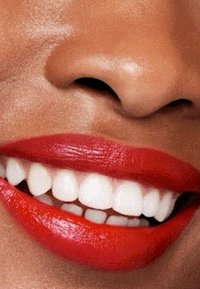 Gros plan sur des lèvres avec un rouge à lèvres rouge vif. Des dents blanches sont visibles et la peau a un teint lisse et chaud. Aucun détail supplémentaire présent.