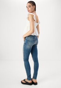 Top peplum blanco con mangas acampanadas, combinado con jeans ajustados azules. La modelo lleva sandalias de plataforma negras, mostrando texturas de tela suaves.