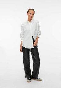 Chemise blanche à manches longues avec manches retroussées, associée à un pantalon large en simili cuir noir et des baskets décontractées avec des accents gris.