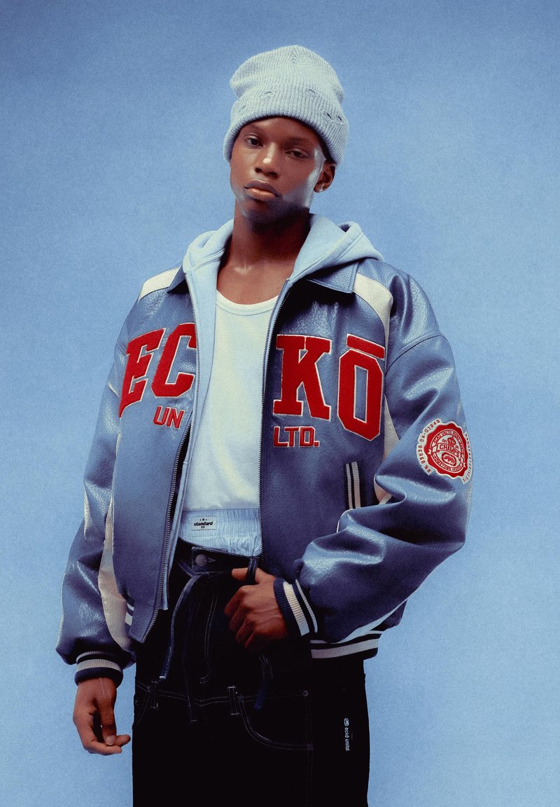 Gray letterman jacket with red "ECKO UNLTD" text, white accents, and a patch. Worn over a white shirt, paired with dark jeans and a light beanie.