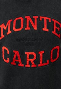 Musta collegepaita, jossa on punaisella brodeerattu teksti "MONTE CARLO" ja "Bjørn Borg 1956" rohkeassa, kaarevassa muotoilussa. Pehmeä rakenne.
