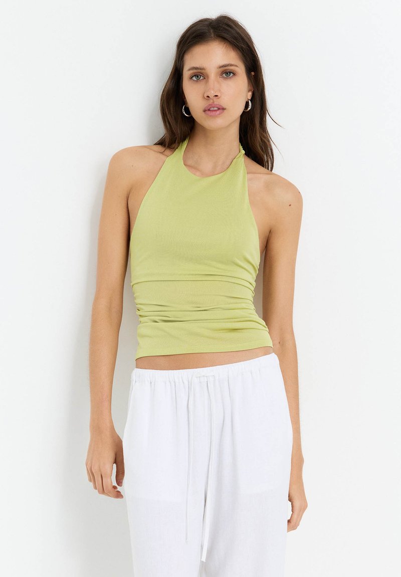 Halter neck top i ljusgrön ribbad tyg, åtsittande design med rynkade detaljer, kombinerad med vita löst sittande byxor.