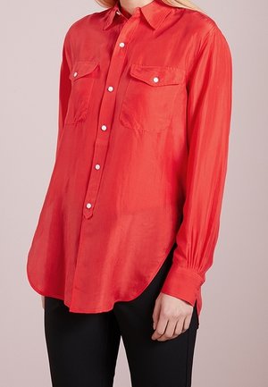Rode blouse met knopen en lange mouwen, met twee borstzakken, gedragen met een zwarte broek bij een staand persoon.