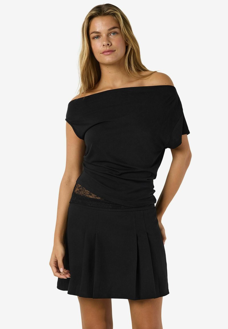 Kobieta w czarnej bluzce bez ramiączek z koronkowym wykończeniem i czarnej spódnicy plisowanej, stojąca na tle białego tła.