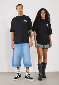 Schwarze oversized T-Shirts mit dem Text "FEEL COOL", kombiniert mit weiten hellen Denim-Shorts und einem Denim-Rock. Schwarze kniehohe Stiefel und Sneakers.
