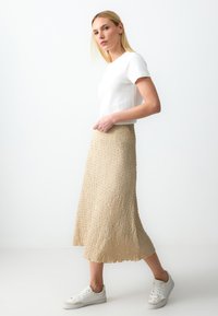 Beige strukturierter Rock mit einem fließenden Design, kombiniert mit einem taillierten weißen Oberteil und weißen Sneakers, vor einem hellen Hintergrund.