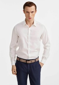 Biała koszula z długim rękawem z zapięciem na guziki, połączona z granatowymi spodniami i jasnobrązowym paskiem. Koszula ma subtelne logo na piersi.