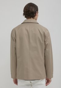 Giacca leggera color sabbia con un design leggermente strutturato, colletto a punte e maniche lunghe; texture liscia, senza motivi visibili né hardware.