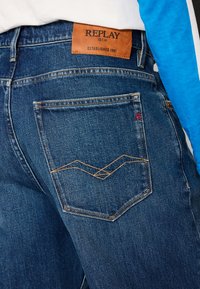 Jean en denim bleu foncé avec un design de poche arrière distinctif, des coutures en zigzag et une étiquette de marque en cuir brun.